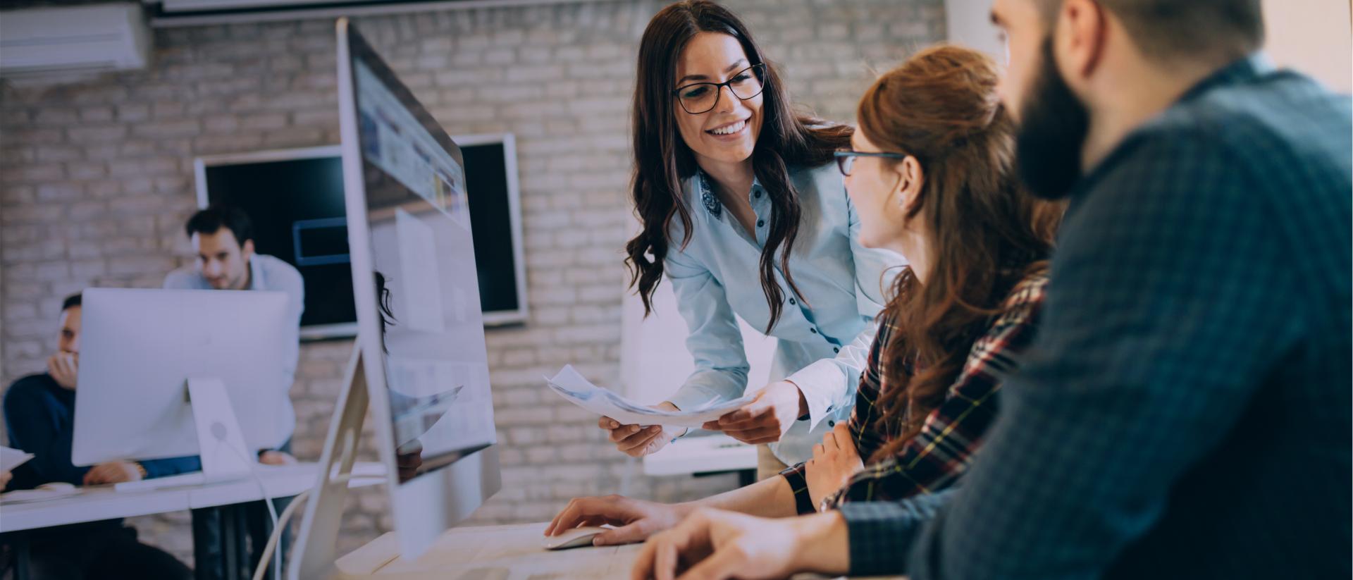 In a work environment, a smiling woman wearing glasses brings documents to 2 other people sitting in front of a computer