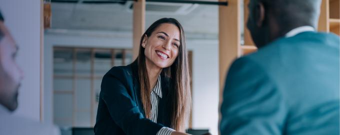 a smiling businesswoman with long brown hair and a document in her hand shakes hands with a man