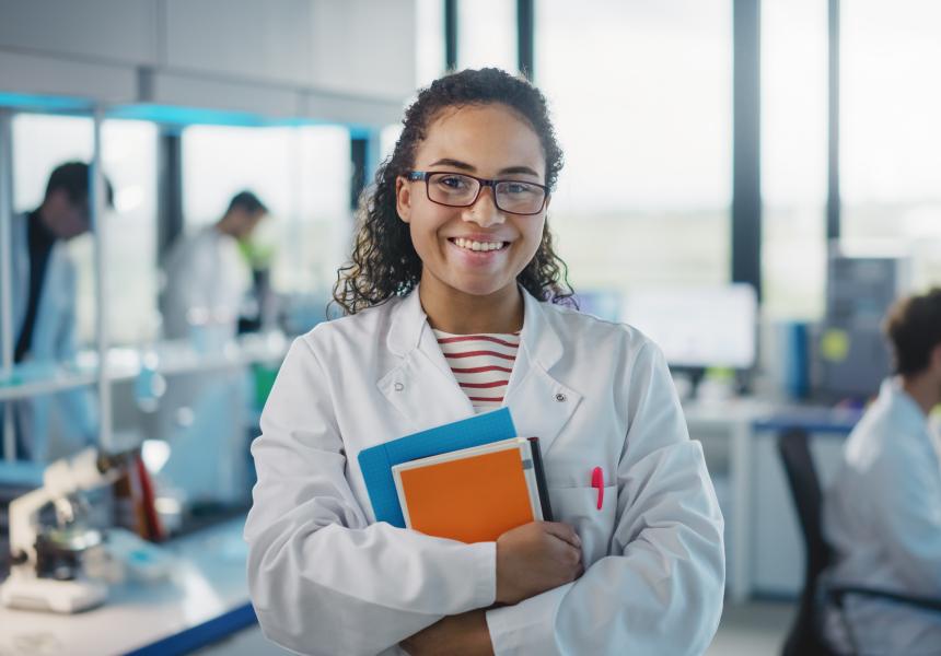 a smiling woman wearing glasses and a white lab coat, holding books in a laboratory