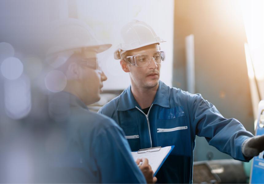 2 people, with helmet and goggles, work at a machine in a factory