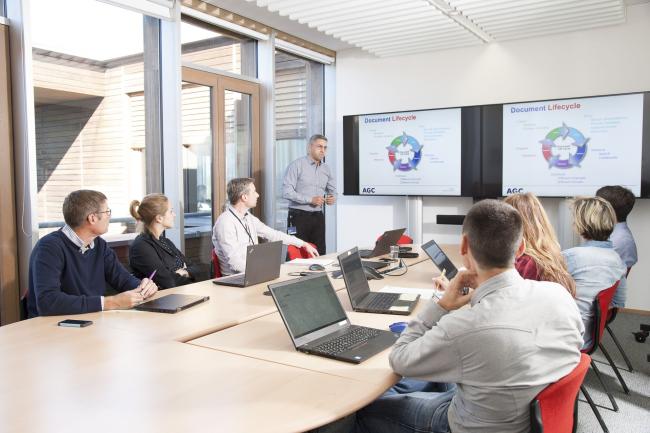 7 people sit in a meeting room and 1 person presents graphics on 2 large screens