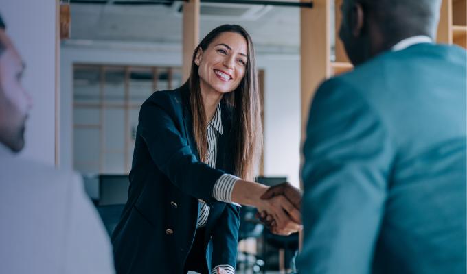 a smiling businesswoman with long brown hair and a document in her hand shakes hands with a man